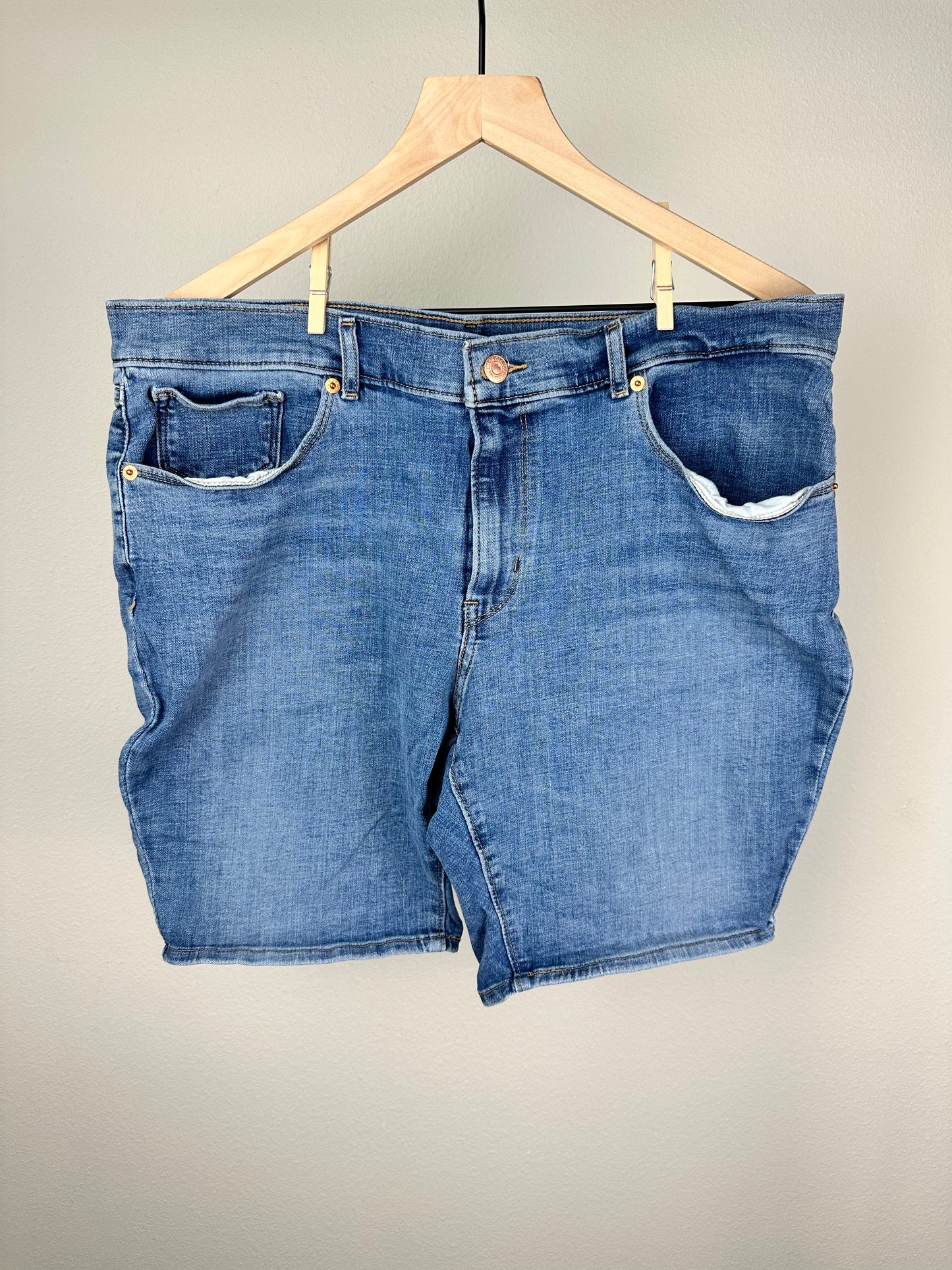 A pair of blue Levi Strauss Bermuda shorts hung on a wooden hanger against a plain background.